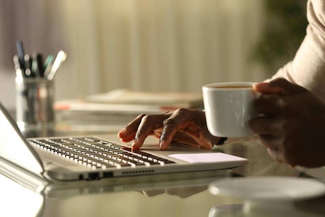 Foto einer Person, die mit einer Kaffetasse in der Hand an einem Notebook arbeitet.