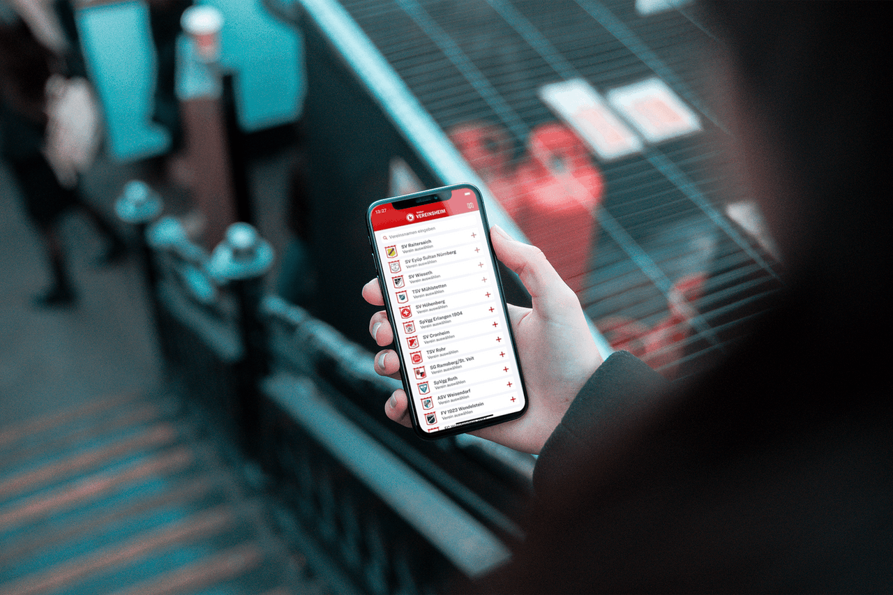 Eine Aufnahme einer Person über die Schulter, die ein iPhone mit der Vereinsheim-App von kicker hält.